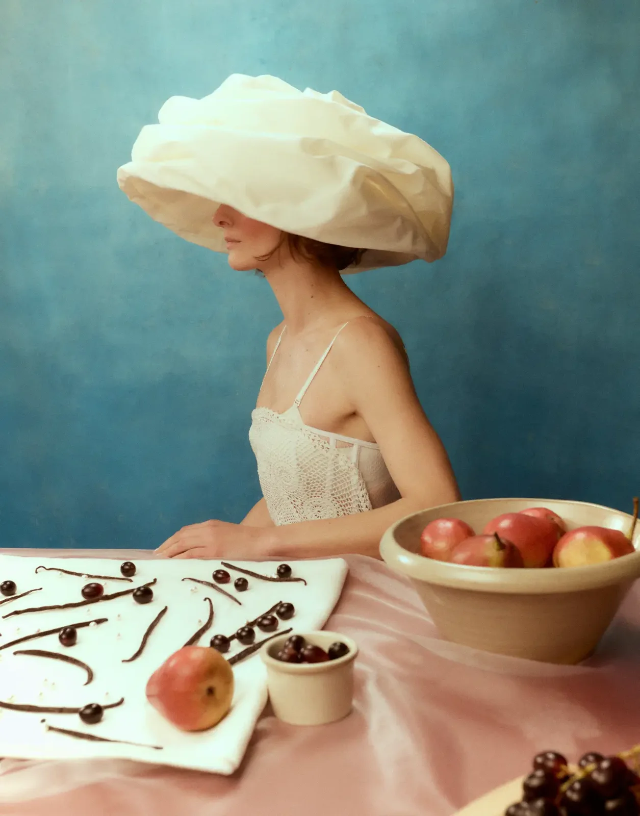 Sombrero escultórico y vestido claro en una mesa con frutas — estilismo de Manel Guisado